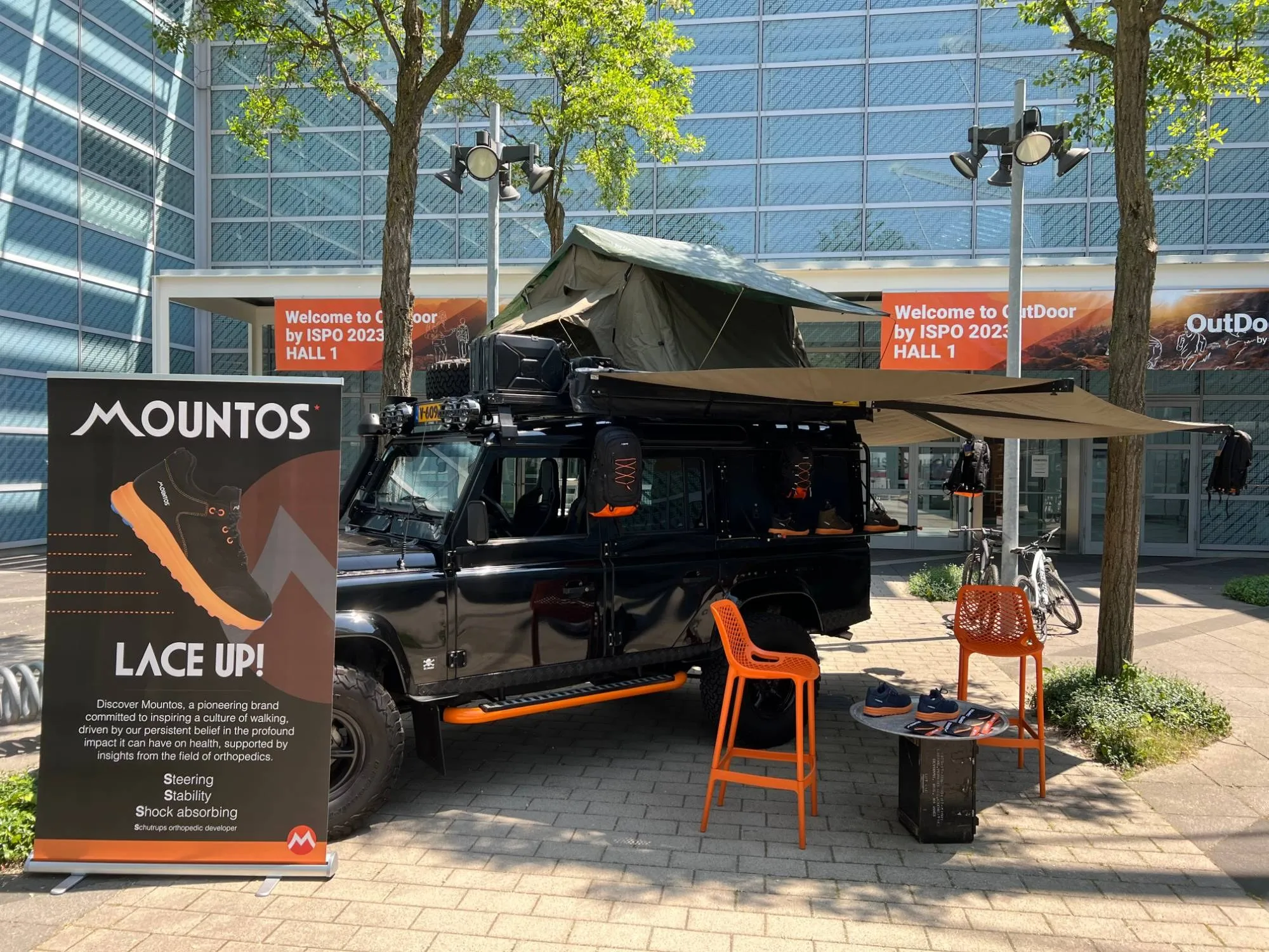Zwarte Land Rover Defender met daktent en een Mountos LACE UP!-banner buiten de beurshal van OutDoor by ISPO 2023