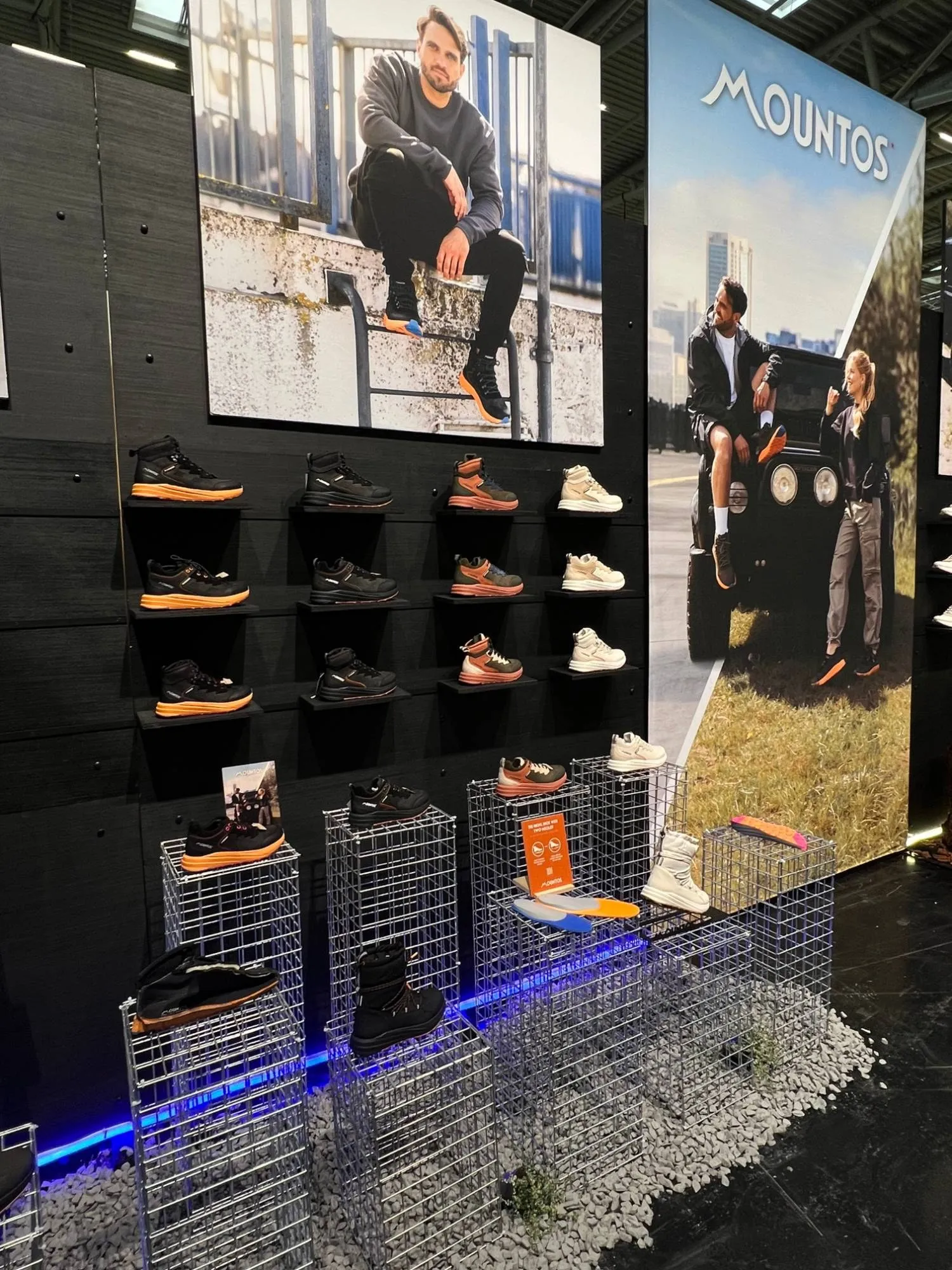 Urban kant van de Mountos-stand: schoenen op zwarte wand boven stalen gaaskorven met blauw licht